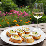 A close-up of a plate of small crostini topped with herbed goat cheese and white wine jelly, with a glass of white wine beside it, set against a blurred garden backdrop with colorful flowers.