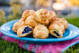A blue plate full of Clif Family Blueberry Jam Filled Gougères sits on a pink cloth napkin on green grass. There are slight yellow flowers in the background.