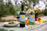 A bottle each of red and white wine on a light wooden table with florals and small plate of fresh food