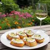 A close-up of a plate of small crostini topped with herbed goat cheese and white wine jelly, with a glass of white wine beside it, set against a blurred garden backdrop with colorful flowers.