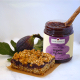 A Figgy Cobbler Bar sits on a square piece of parchment paper with a fresh fig plucked from a tree sitting next to an open jar of Clif Family Fig Jam.