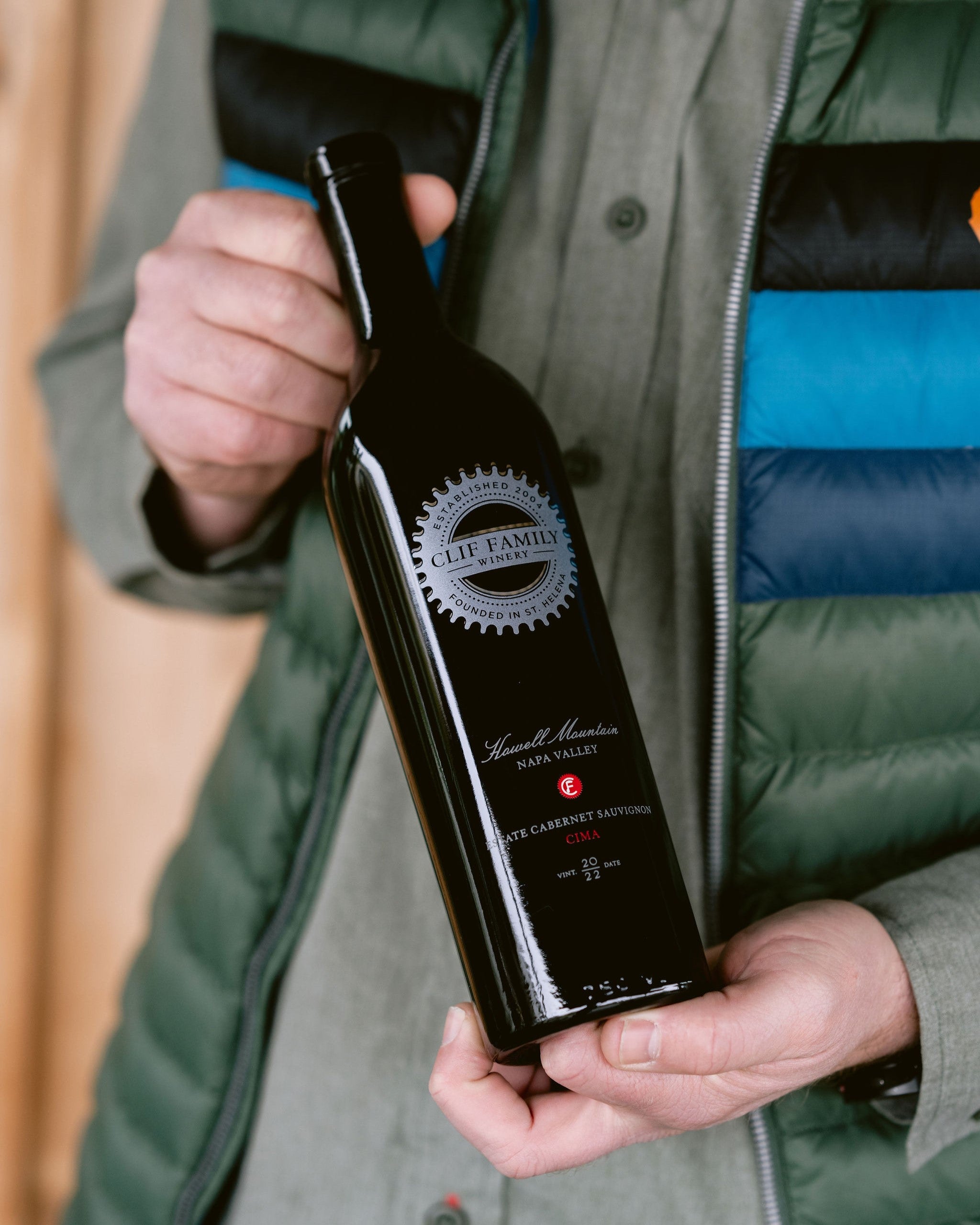 Person holding a Clif Family Cima Howell Mountain Cabernet Sauvignon bottle in a green and blue jacket.
