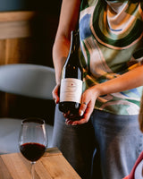Person holding a bottle of Clif Family Grenache with a glass on a wooden table.