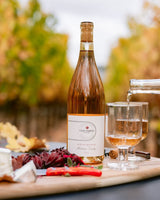 Clif Family Rosé of Grenache bottle with a cork, a glass of wine, and a charcuterie board with cheese and dried herbs on a blurred outdoor background.