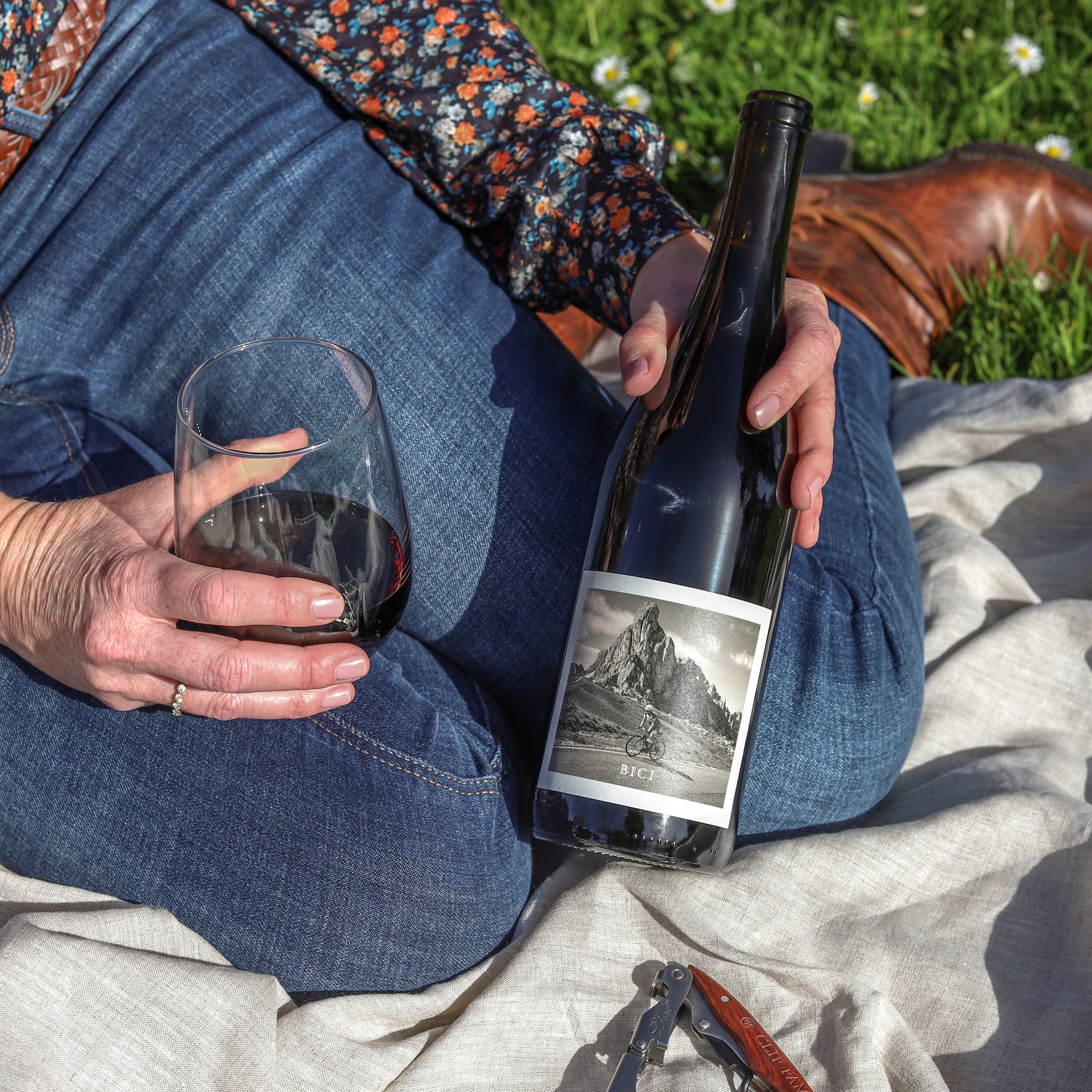 Woman in boots sitting on a picnic blanket with a bottle of wine in one hand and glass of the wine in her other hand