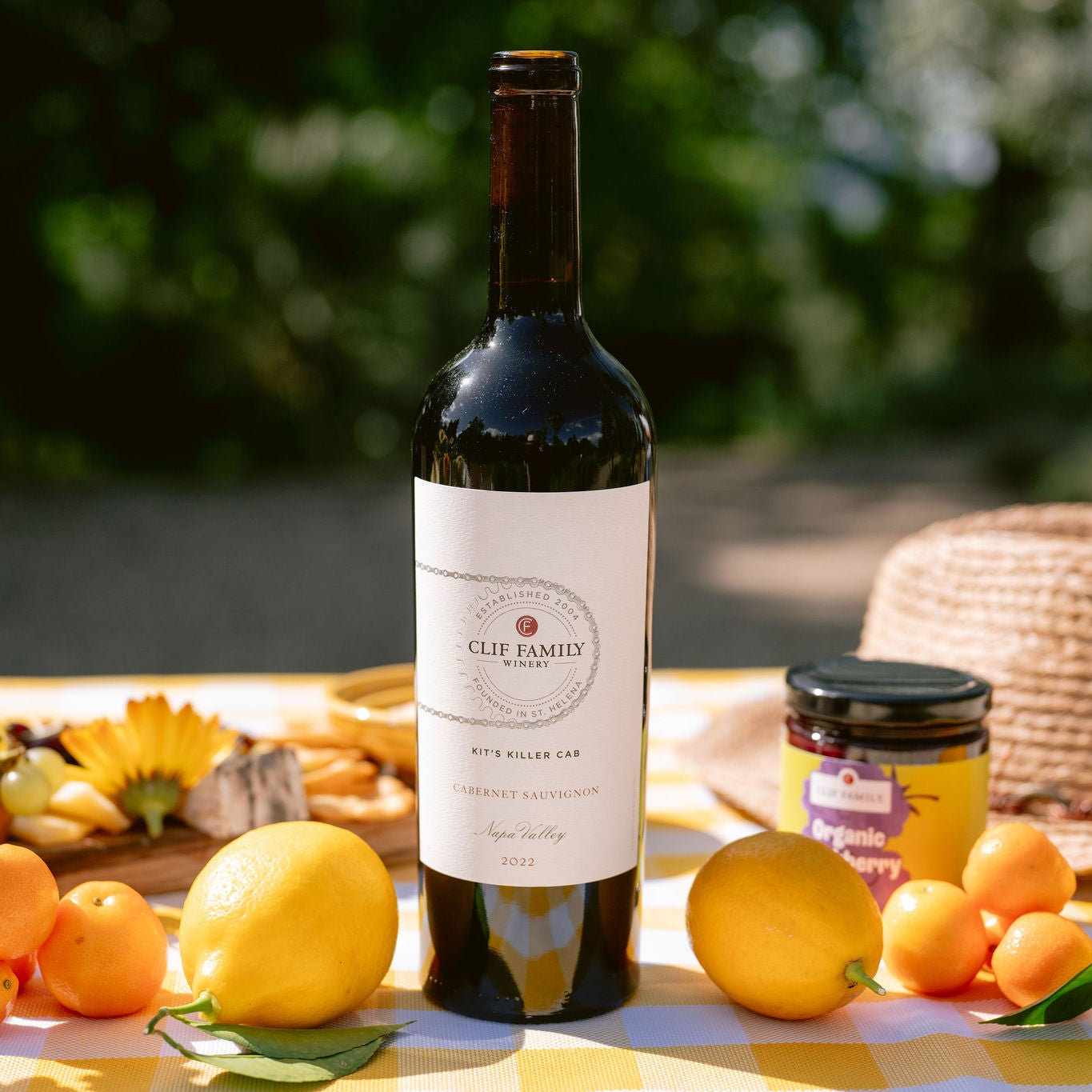 A bottle of Clif Family Kit's Killer Cab is displayed on a yellow and white checkered picnic blanket outdoors, surrounded by lemons, oranges, a straw hat, and a jar of honey.