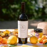 A bottle of Clif Family Kit's Killer Cab is displayed on a yellow and white checkered picnic blanket outdoors, surrounded by lemons, oranges, a straw hat, and a jar of honey.