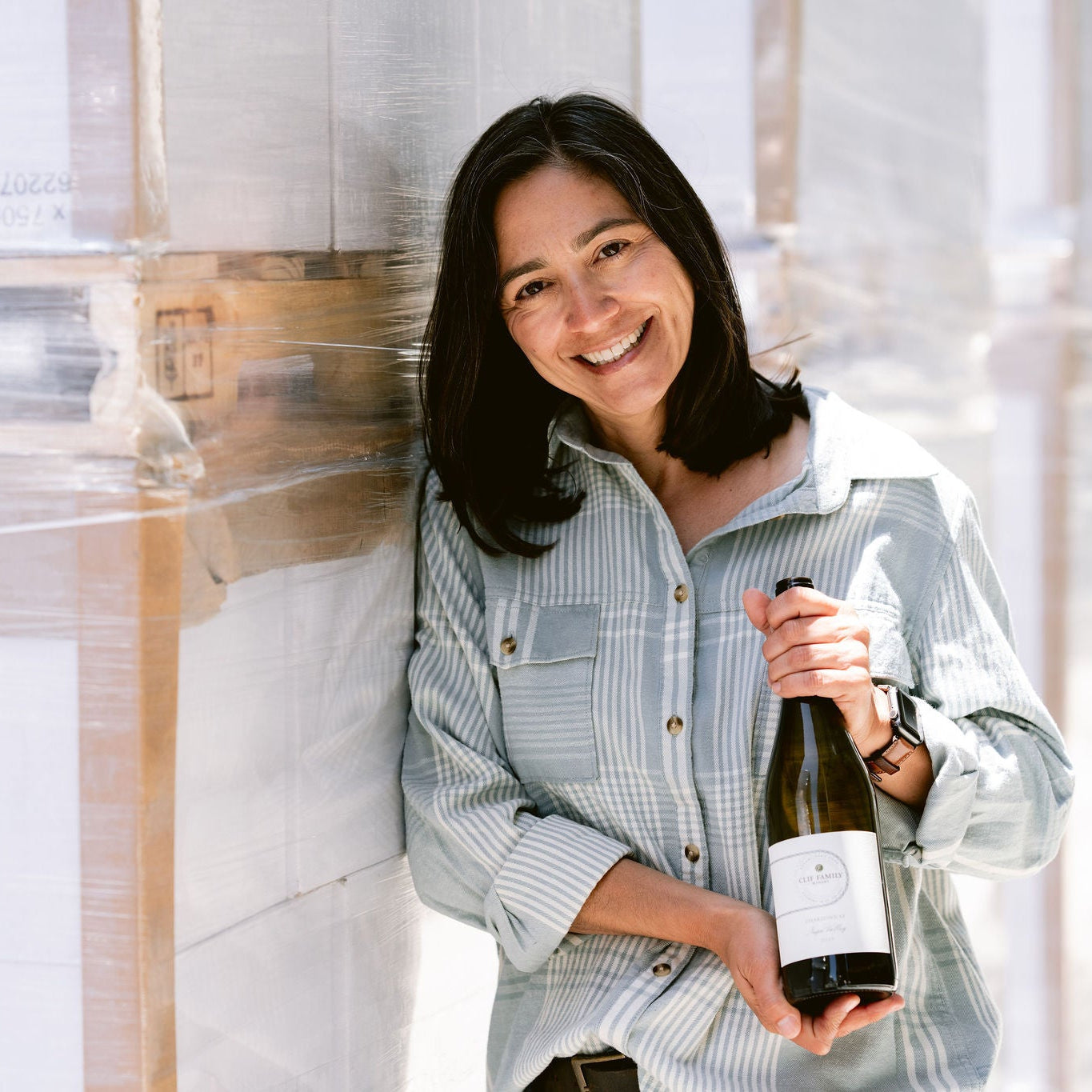 Winemaker Laura Barrett Holding a bottle of 2023 Chardonnay in front of pallets of wine