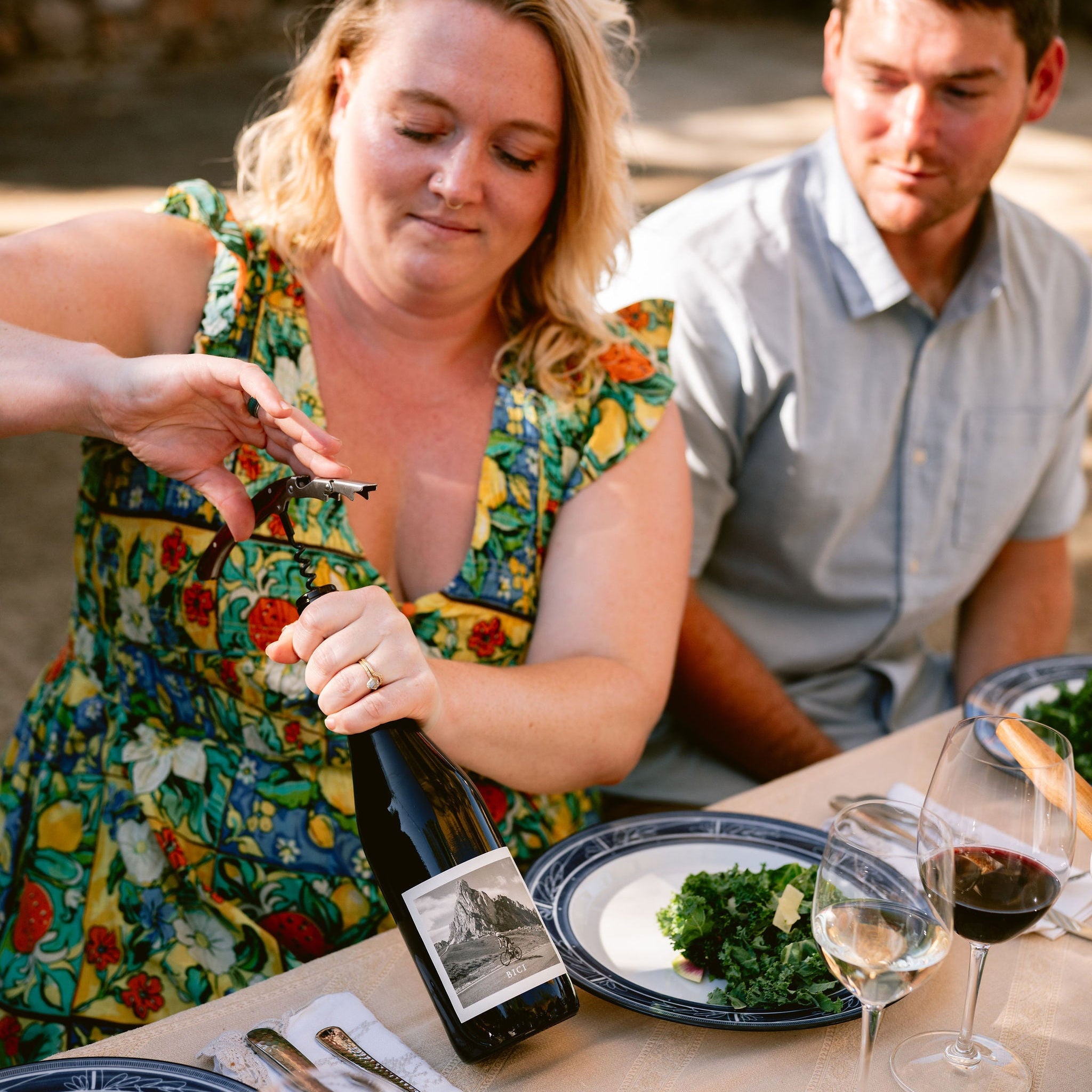 A woman in a colorful floral dress opens a bottle of red wine at a dining table, with a man seated beside her and plates of food and wine glasses on the table.
