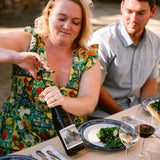 A woman in a colorful floral dress opens a bottle of red wine at a dining table, with a man seated beside her and plates of food and wine glasses on the table.
