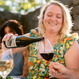 A woman in a colorful floral dress smiles while pouring red wine from a dark bottle into a glass, with another person holding a glass of white wine in the foreground and a stone wall in the background.