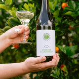 Person holding a bottle of wine and a glass with a green leafy background