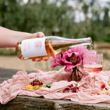 A hand pours pink wine from a bottle with a white label into a glass, set on a picnic blanket outdoors with flowers, fruit, and cheese.