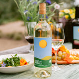 A bottle of white wine with a light blue and green label stands on a table outdoors, surrounded by wine glasses, a salad, grapes, and flowers in a blurred garden setting.