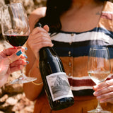 A person in a striped top holds a dark wine bottle with a mountain label while two hands raise wine glasses in a sunny outdoor setting with a stone wall background.