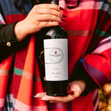 Person holding a bottle of wine with a visible label, wearing a colorful plaid garment.