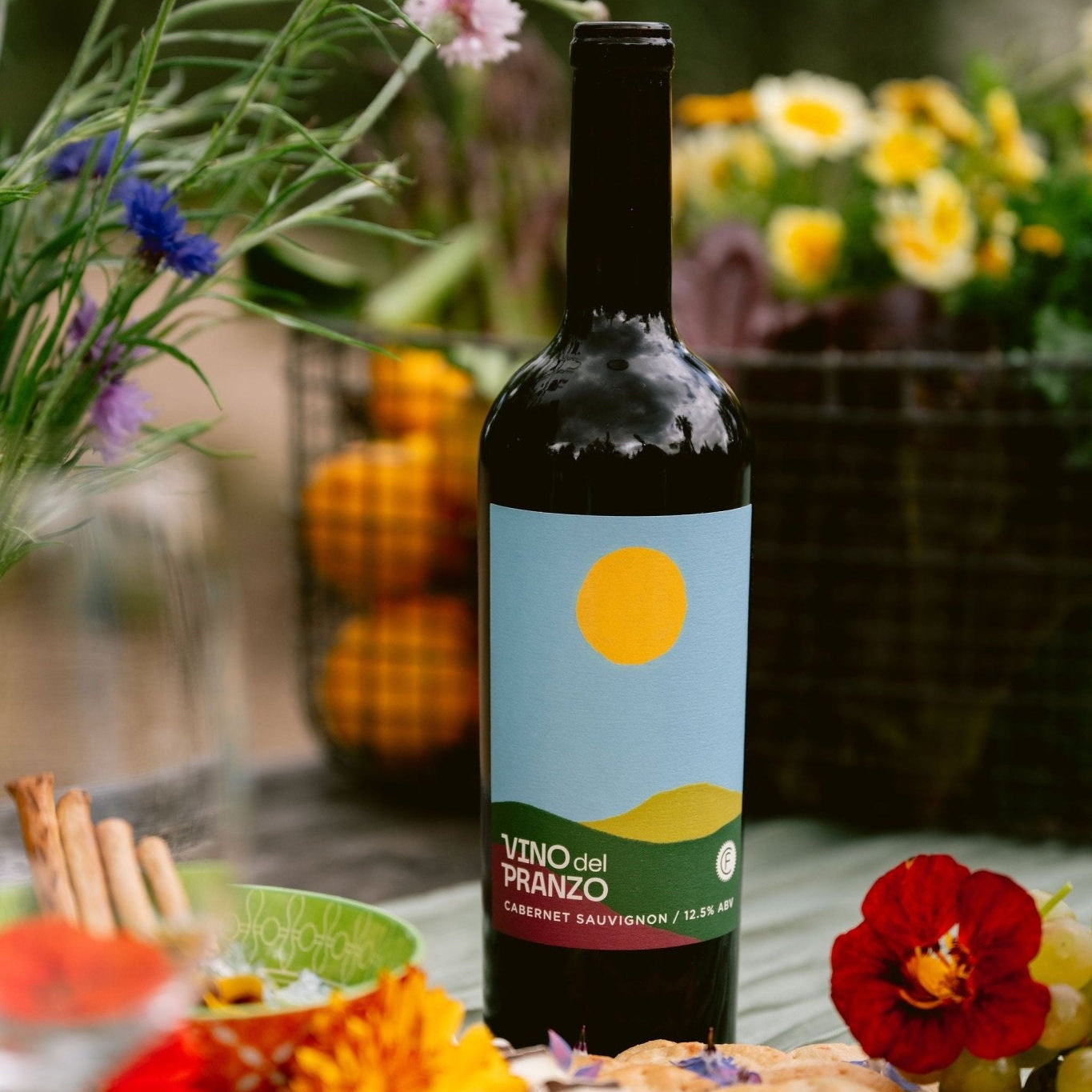 A bottle of Vino del Panzo Cabernet Sauvignon wine stands on a table surrounded by flowers, bread, grapes, and fruit in an outdoor garden setting.